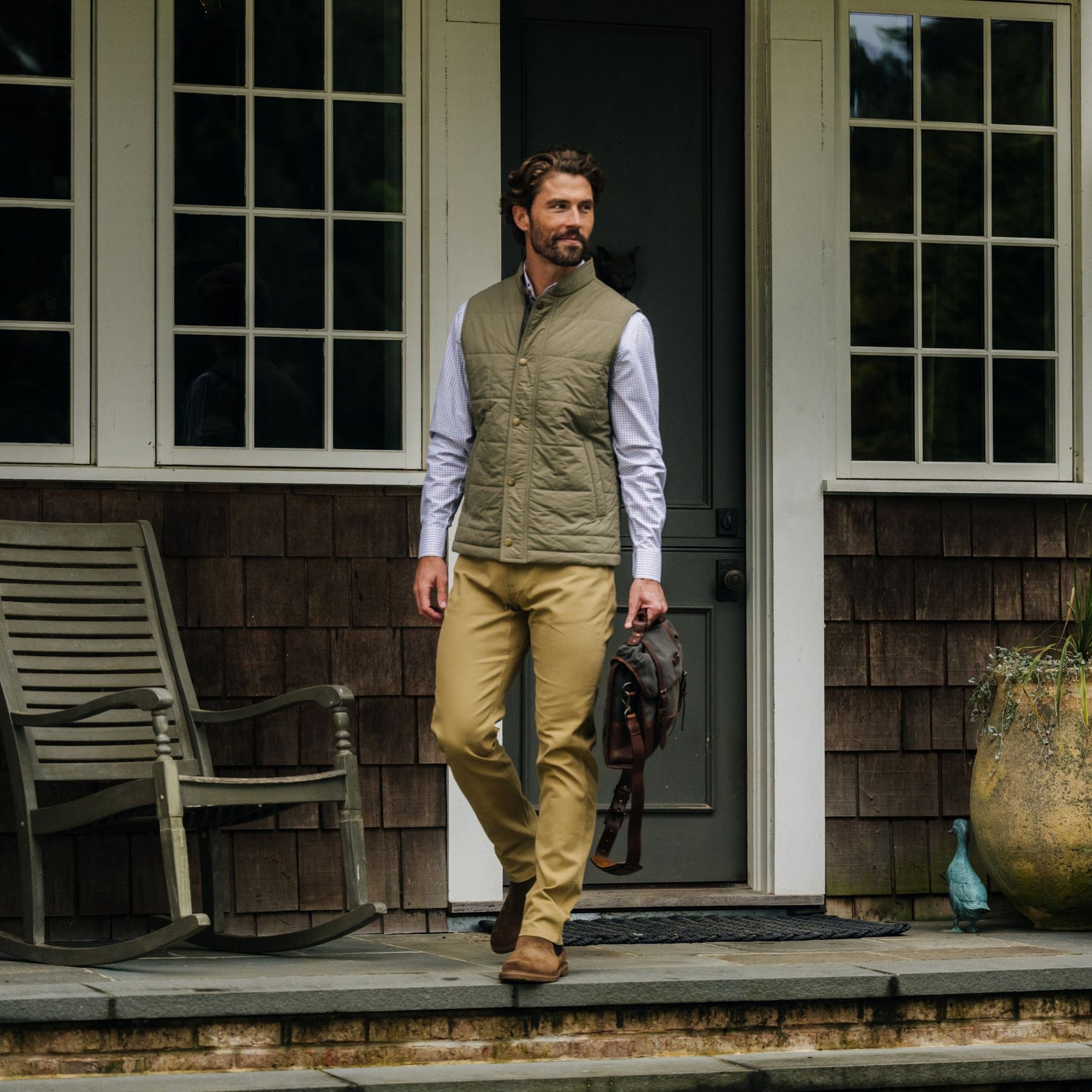 Man in a Tom Beckbe Burgess Quilted Vest and khaki pants stands on a porch with a brown bag near a rocking chair.
