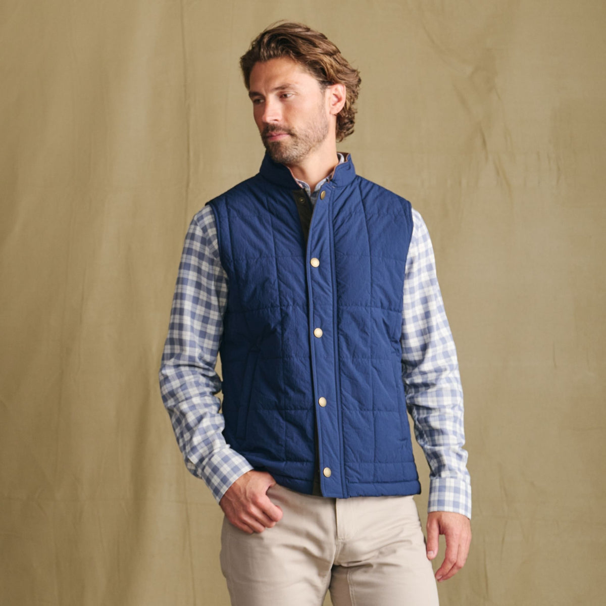 Man in Tom Beckbe Burgess Quilted Vest over plaid shirt, beige pants, stands before a plain tan background.
