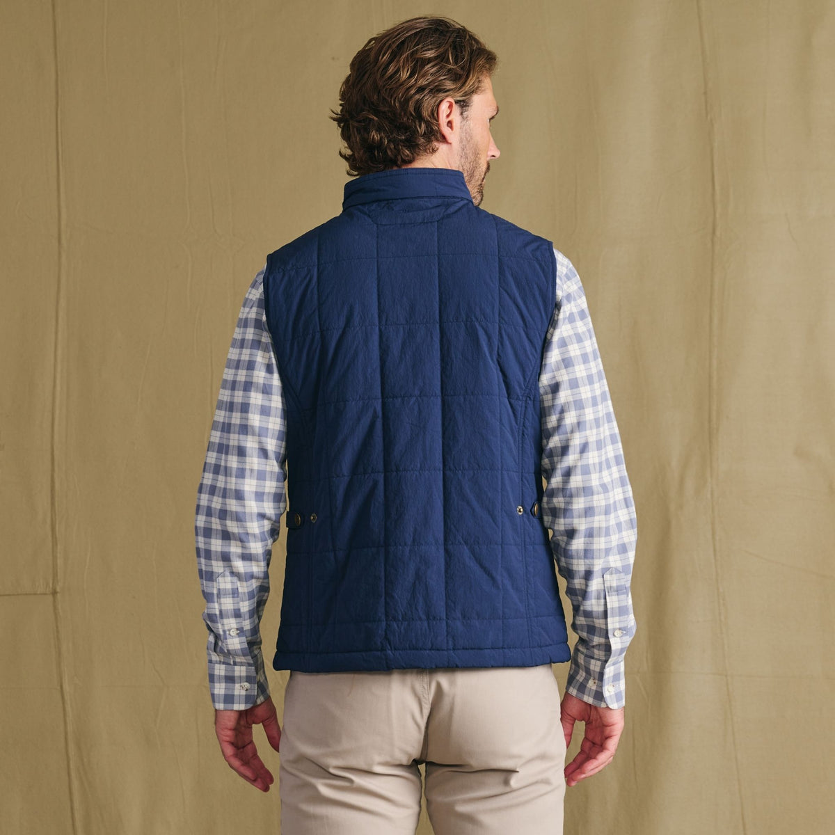 Man facing away in Tom Beckbe Burgess Quilted Vest over plaid shirt and beige pants, standing before a tan background.