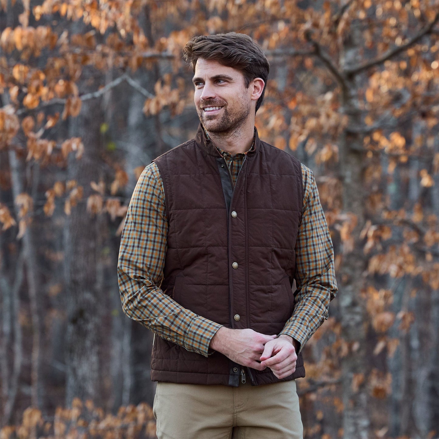 Man wears Tom Beckbe Burgess Quilted Vest over plaid shirt outdoors with autumn trees and brown leaves in the background.