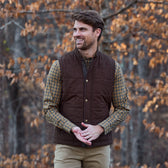 A man wears the Tom Beckbe Burgess Quilted Vest over a plaid shirt and khakis, surrounded by autumn leaves.