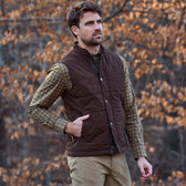 A man in a Tom Beckbe Burgess Quilted Vest, plaid shirt, and khaki pants stands outdoors amid autumn leaves.
