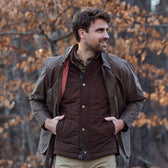 Man outdoors in a Tom Beckbe Burgess Quilted Vest and jacket, hands in pockets, autumn foliage behind him.