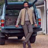 Man in a Tom Beckbe Burgess Quilted Vest and brown pants leans on an SUV trunk filled with travel bags.