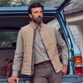 A man in a Tom Beckbe Burgess Quilted Vest and plaid shirt leans on an open blue SUV trunk packed with bags.