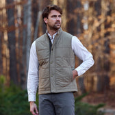 A man wears the Tom Beckbe Burgess Quilted Vest over a button-down shirt, standing outdoors amid autumn foliage.