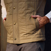 Person in a tan Tom Beckbe Burgess Quilted Vest, hands in pockets, with gray pants and a white checkered shirt.