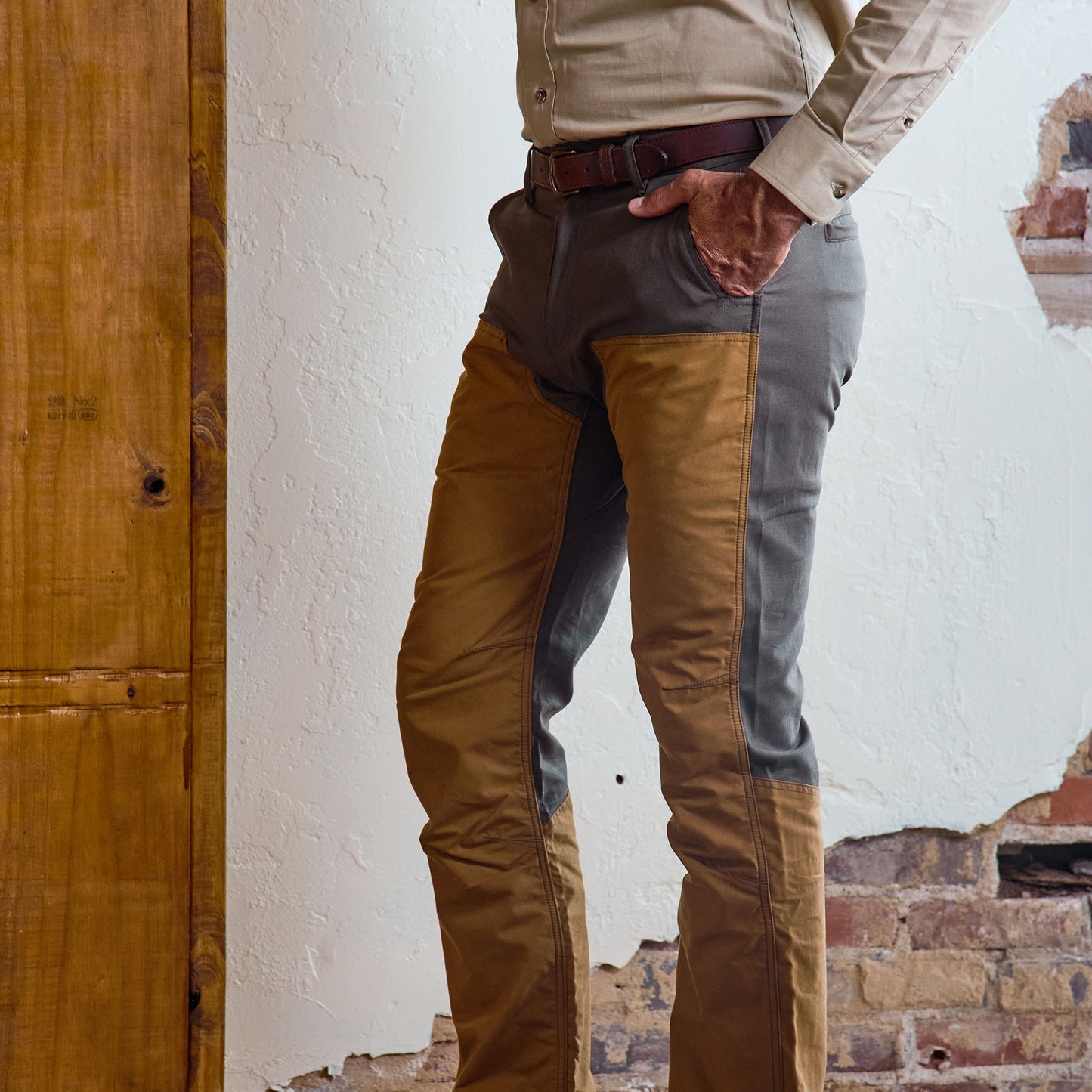 A man in Tom Beckbe’s Burwell Brush Pant and long-sleeve shirt stands by a textured, exposed brick wall.