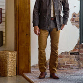 A man indoors wears Tom Beckbe’s Burwell Brush Pant with a dark jacket and boots beside a textured wall and wooden panel.