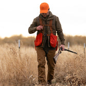 A person in an orange cap and vest walks through tall grass wearing Tom Beckbe Burwell Brush Pants, carrying a shotgun.