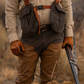 Wearing Tom Beckbe Burwell Brush Pants and gloves, a person stands in dry grass holding a shotgun with two chest pouches.