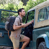 Man with a Tom Beckbe Canvas Backpack climbing into a vintage off-road vehicle in the outdoors.