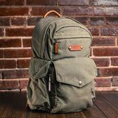 Tom Beckbes Canvas Backpack, green waxed canvas with laptop sleeve and leather handle, set against a brick wall.