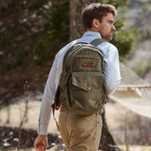 Bearded man in a white shirt with a Tom Beckbe canvas backpack stands outdoors by trees and a hammock.