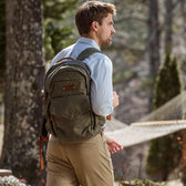 A man outdoors with a Tom Beckbe Canvas Backpack wears a light blue shirt, khaki pants, with a hammock in the background.