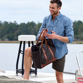 Man in a blue plaid shirt and shorts stands on a boat with Tom Beckbes Canvas Carryall Bag, lake and trees behind.