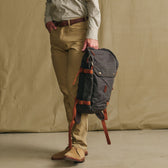 A person in khakis holds a Tom Beckbe Canvas Daypack with brown straps against a beige backdrop.