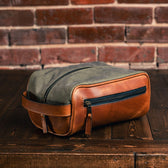 The Canvas Dopp Kit by Tom Beckbe, a brown and green waxed canvas toiletry bag, sits on wood against a brick wall.
