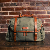 A Canvas Field Bag by Tom Beckbe in green and brown leather rests on wood, showcasing its rugged utility against a brick backdrop.