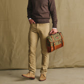 A person in a brown sweater holds the Tom Beckbe Messenger Bag with a leather handle against a tan background.