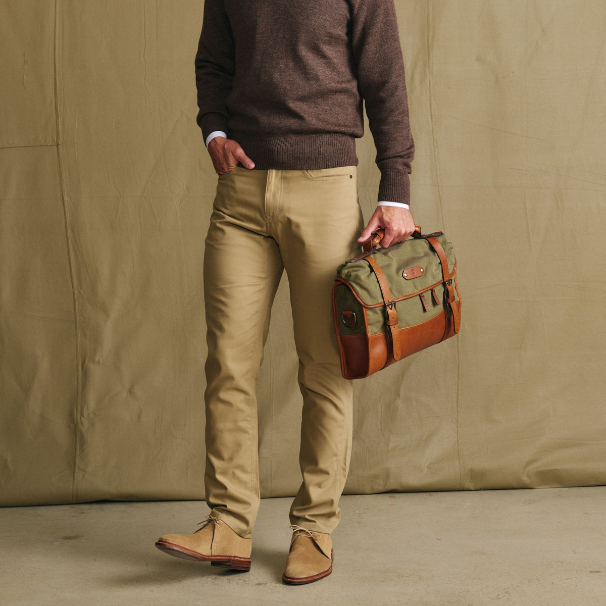 A person in a brown sweater holds the Tom Beckbe Messenger Bag with a leather handle against a tan background.