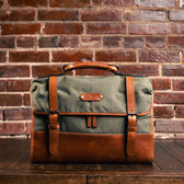 A Tom Beckbe Messenger Bag in green and brown leather with a laptop sleeve sits on a wooden table against a brick wall.