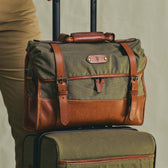 The Tom Beckbe Messenger Bag in olive green and brown rests on a matching suitcase, both featuring handles and straps.