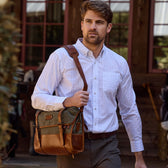 A man in a white shirt carries a Tom Beckbe Messenger Bag outside a wooden building with red window frames.