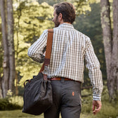 A man wearing a checkered shirt and gray pants walks in a forest, carrying a Tom Beckbe Canvas Tailgater Bag.