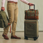 A man holds a green backpack and rolls a Tom Beckbe Canvas Wheeled Carry-On Bag on a light floor.