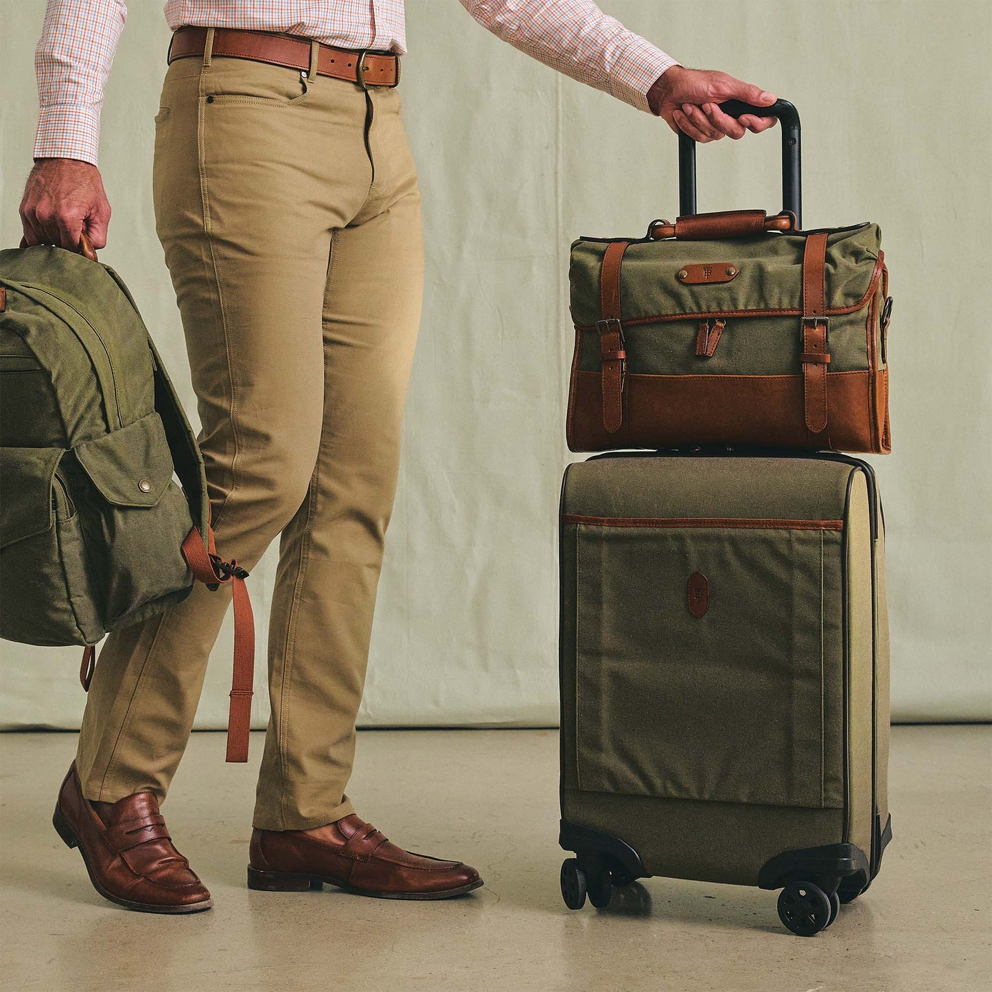 A man holds a green backpack and rolls a Tom Beckbe Canvas Wheeled Carry-On Bag on a light floor.