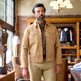 A man in a Tom Beckbe Chatom Jacket and khaki pants stands in a stylish room with brick walls and large windows.