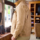 Man wears a Tom Beckbe Chatom Jacket indoors by a window with shelves, chairs, and bags in the background.