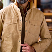 Man in Tom Beckbe Chatom Jacket, tan waxed, over brown shirt, buttoning up with one hand; face partly visible.