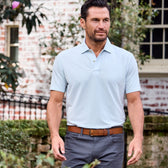 A man in a classic Coastal Polo Shirt by Tom Beckbe and gray pants strolls outside near a brick building with plants.