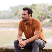 Man sitting outdoors on a brick ledge wearing a Tom Beckbe Coastal Polo Shirt in orange and gray pants, gazing thoughtfully.