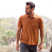Man in a Coastal Polo Shirt by Tom Beckbe and dark pants walking outdoors with trees in the background.