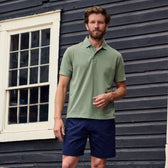 A man in a classic green Coastal Polo Shirt by Tom Beckbe and navy shorts stands before a wooden building with white-framed windows.