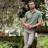 A man in a Tom Beckbe Coastal Polo Shirt and beige pants leans on a mossy stone wall in a lush garden.