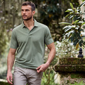 A man in a Tom Beckbe Coastal Polo Shirt and khaki pants walks by a mossy stone wall surrounded by lush greenery.