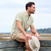 Man in a Tom Beckbe Coastal Polo Shirt and khaki shorts sits on a wooden fence, holding a hat, with nature behind.
