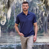 A man in a Coastal Polo Shirt by Tom Beckbe strolls near water, framed by trees draped with hanging moss.