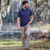Man in a Tom Beckbe Coastal Polo Shirt and beige pants walking on grass near a lake with trees in the background.