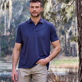 A person in a navy Tom Beckbe Coastal Polo Shirt and beige pants stands outdoors near trees, a pond, and moss.
