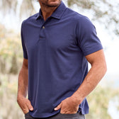 A man in a Tom Beckbe Coastal Polo Shirt stands with hands in pockets, blurred greenery and trees behind.