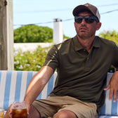 A man in sunglasses and a cap relaxes outdoors on a striped bench, holding a drink, wearing a Tom Beckbe Coastal Polo Shirt.