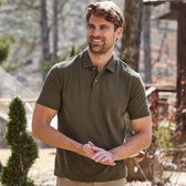A man in a green Coastal Polo Shirt by Tom Beckbe smiles outdoors, with trees and a wooden structure behind him.