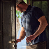 A man in a Tom Beckbe Coastal Polo Shirt opens a door while holding a glass.