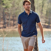 A man in a Tom Beckbe Coastal Polo Shirt and beige shorts walks near a lake with trees in the background.