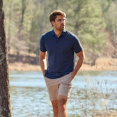 Man in a Tom Beckbe Coastal Polo Shirt and shorts strolling by a lake with trees.